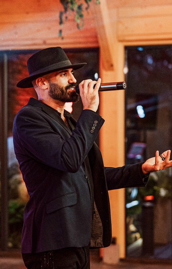 Sami Harb in a black fedora and jacket, singing at an evening event